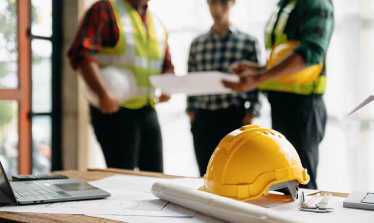 Three construction workers discuss plans in the background as the focus remains on a hard helmet.